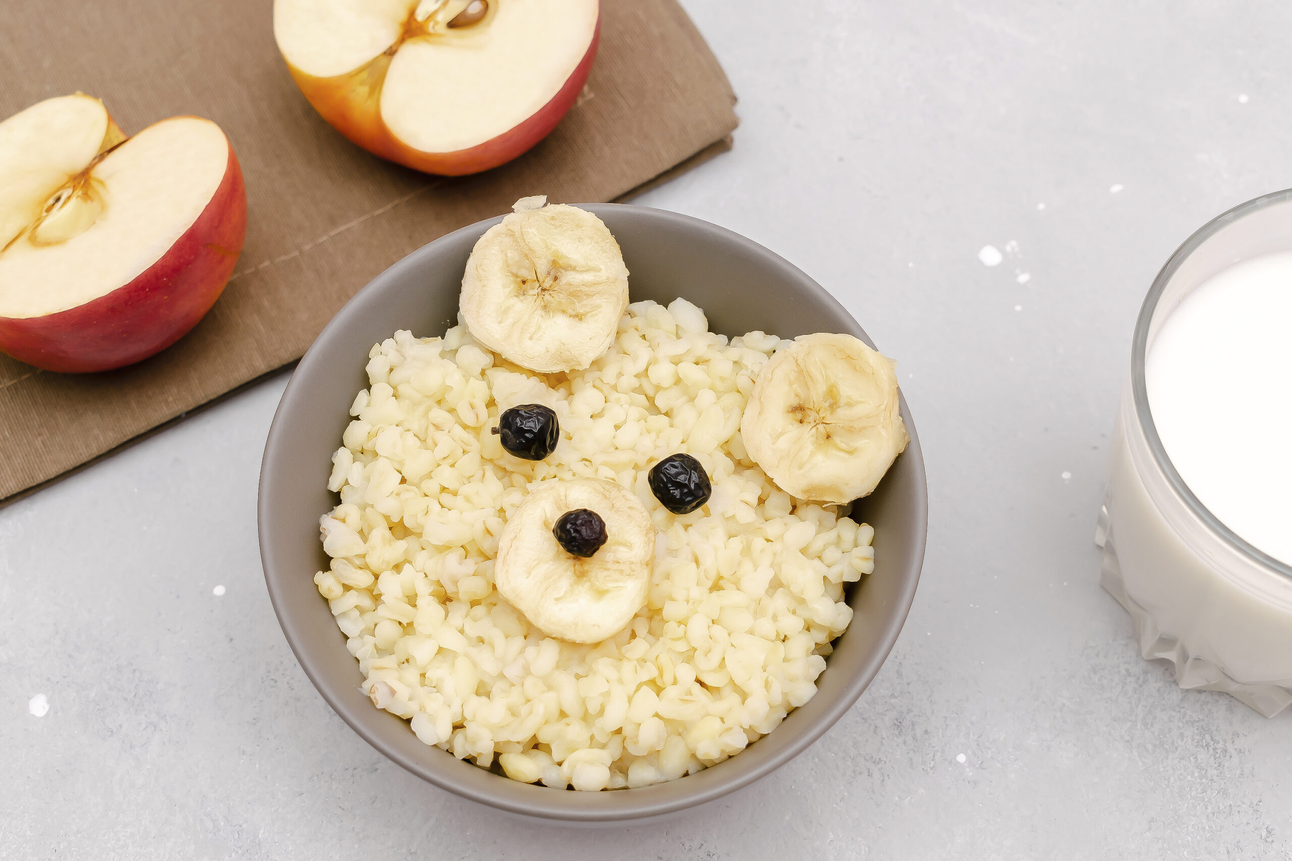 Funny cute kids childrens baby's healthy breakfast lunch oatmeal porridge in bowl look like bear face decorated with apple, banana, dried berry fruits,milk. dessert food art on gray table